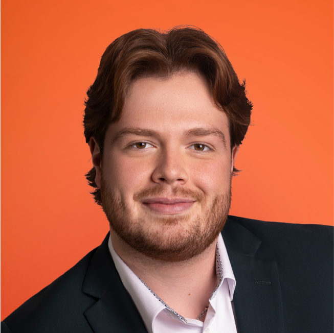 Portrait d'un homme souriant avec des cheveux mi-longs et une barbe, portant un costume foncé et une chemise blanche. L'arrière-plan est d'un orange vif.