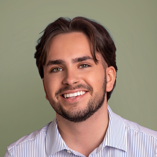 Portrait d'un homme souriant avec des cheveux bruns mi-longs et une barbe, portant une chemise à rayures blanches et bleues. L'arrière-plan est d'un vert doux.