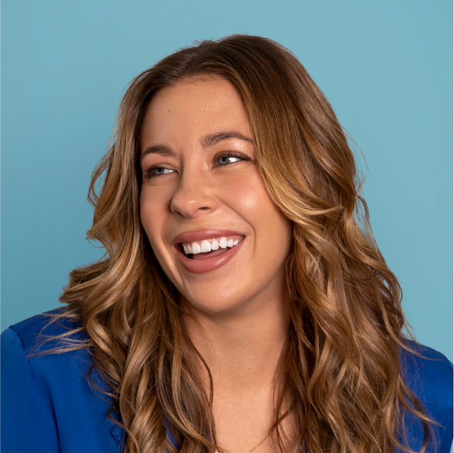 Portrait d'une femme souriante aux cheveux longs et ondulés, portant un blazer bleu. L'arrière-plan est d'un bleu clair.
