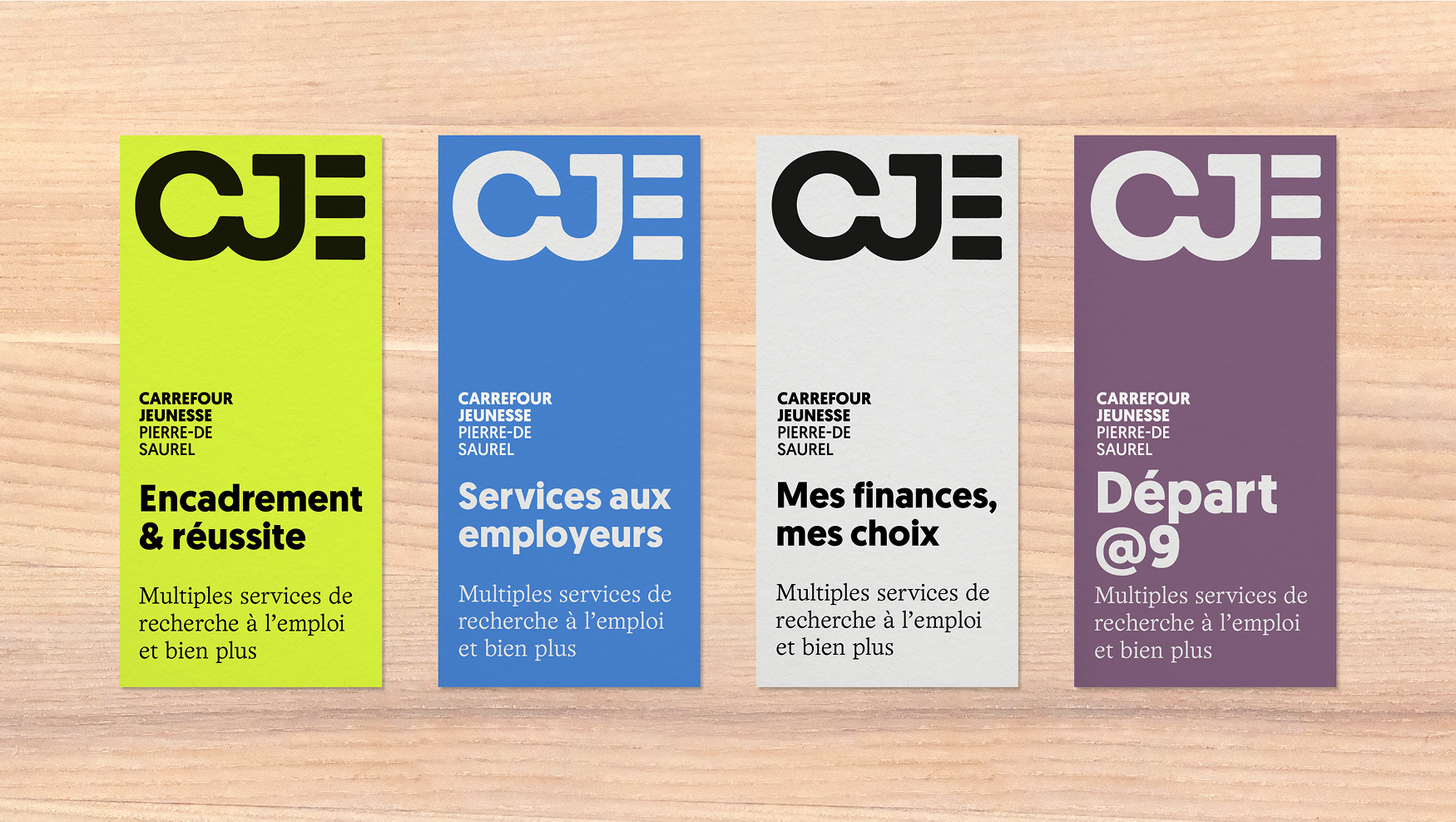 Quatre affiches du Carrefour Jeunesse Pierre-De Saurel alignées sur une surface en bois, chacune avec un fond coloré distinct et un texte différent : "Encadrement & réussite" (vert), "Services aux employeurs" (bleu), "Mes finances, mes choix" (gris), et "Départ @9" (violet).