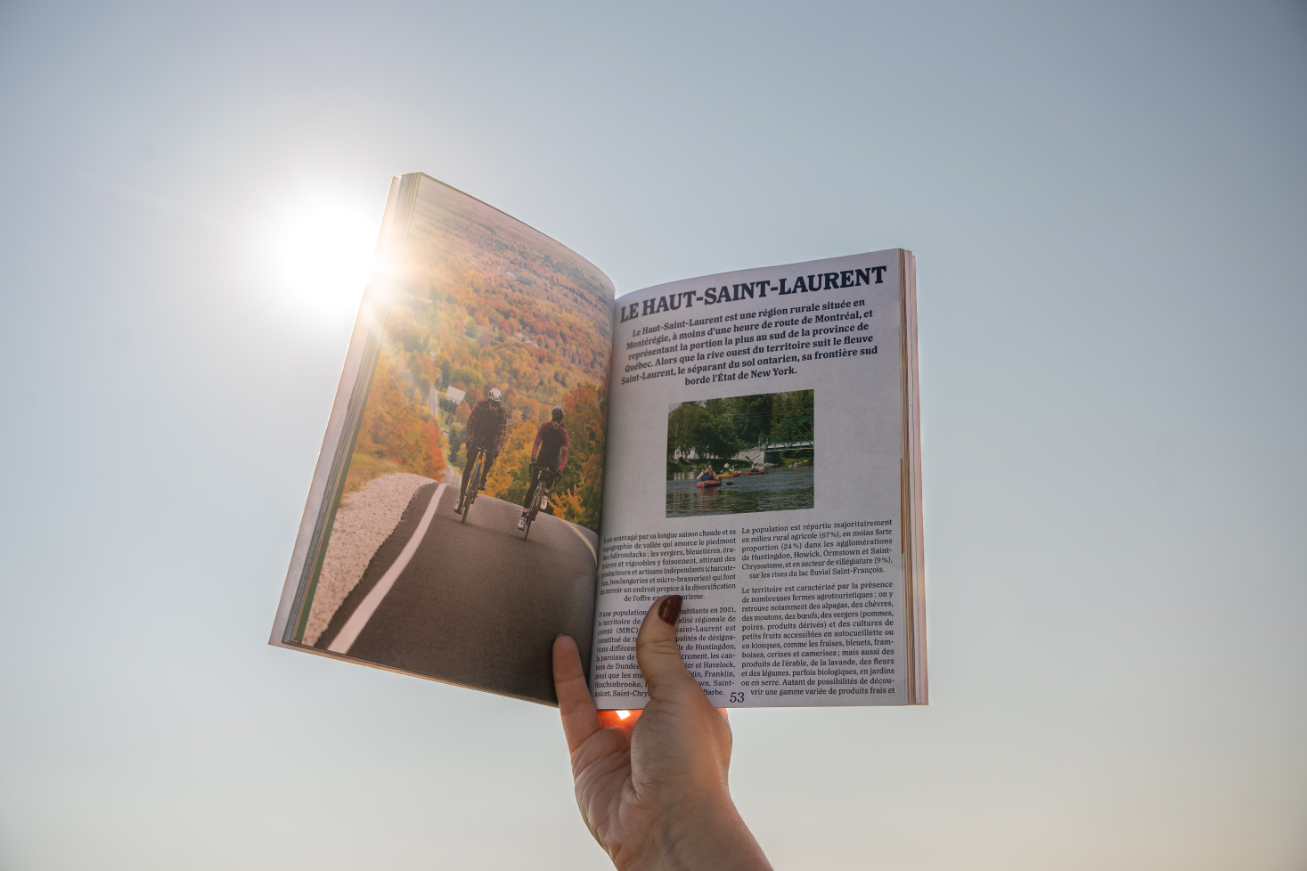 Main tenant un magazine ouvert présentant la région du Haut-Saint-Laurent avec des cyclistes sur une route entourée de paysages automnaux, sous un ciel ensoleillé.
