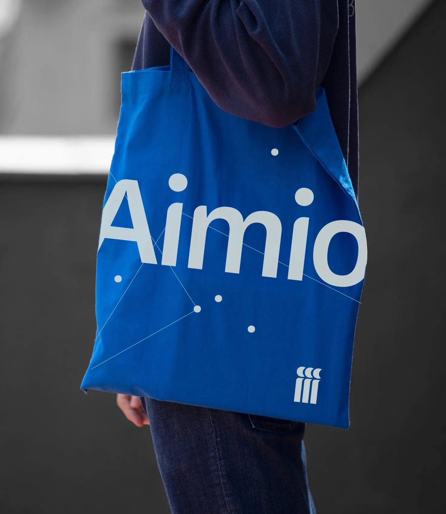 Sac bleu porté à l'épaule, imprimé avec le texte « Aimio » en blanc, des motifs de points reliés par des lignes et un logo minimaliste blanc en bas.
