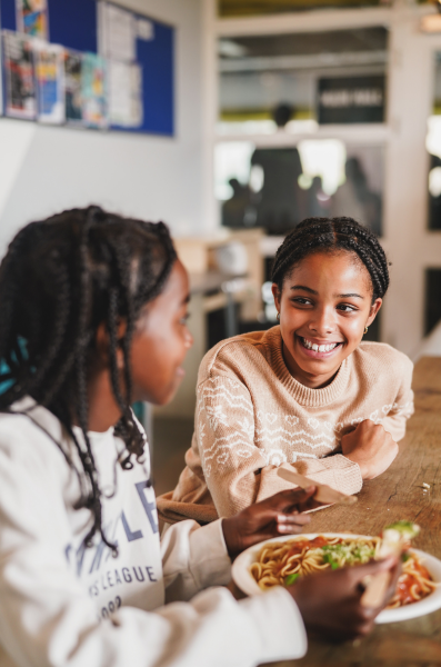 Two young girls chat and eat pasta.