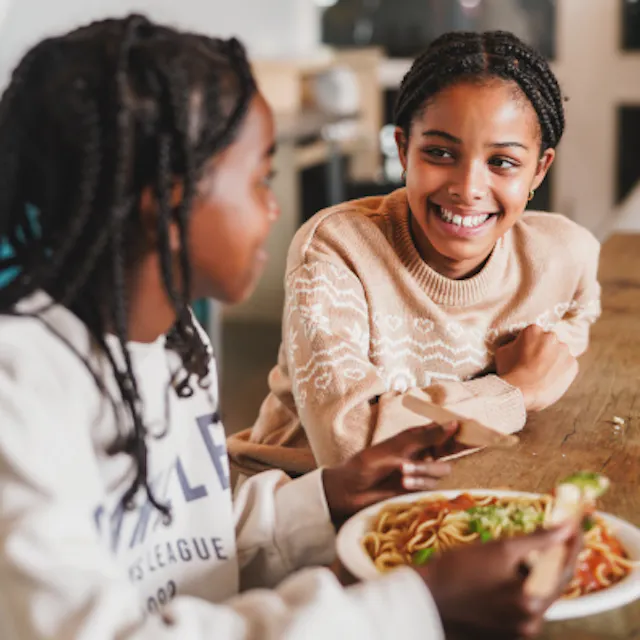Two young girls chat and eat pasta.
