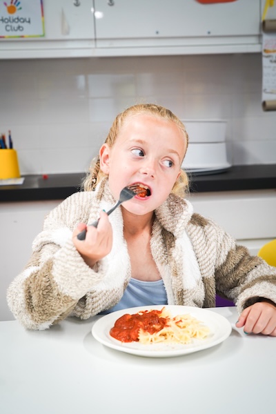 Child eating pasta with tomato sauce at a table, holding a fork in a kitchen setting.