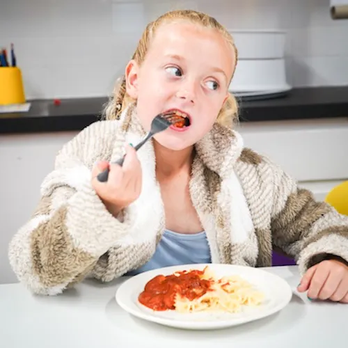 Child eating pasta with tomato sauce at a table, holding a fork in a kitchen setting.