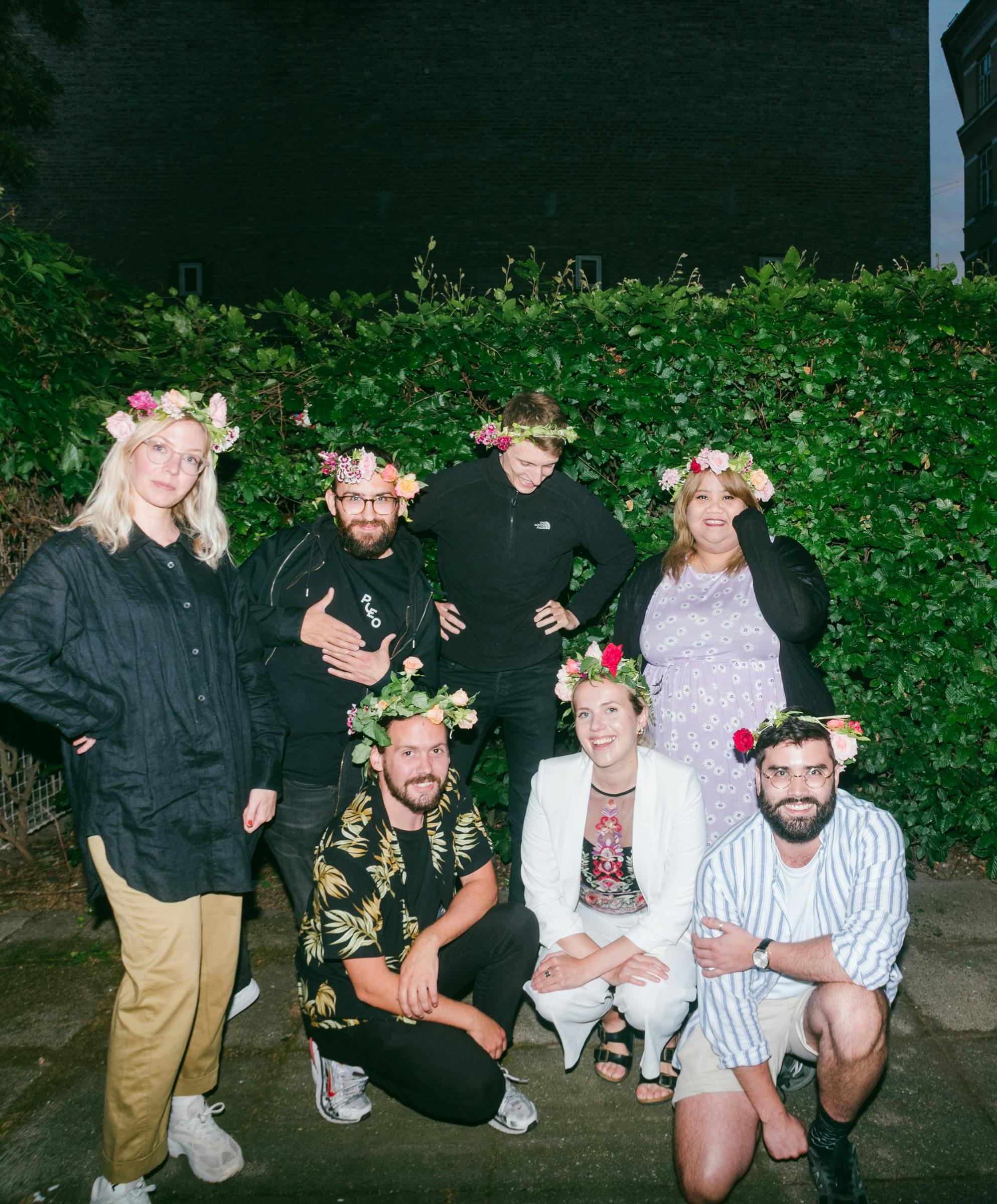 A photograph of seven Pleo employees with four standing and 3 crouched infront all wearing flower crowns and with big smiles