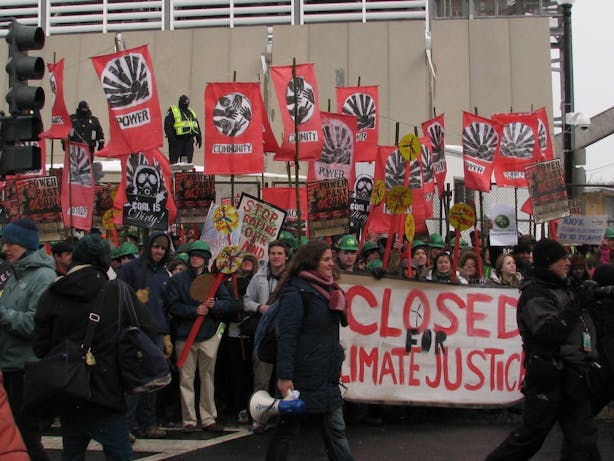 a peaceful demonstration with a group of young people holding signs and banners, there is a woman carrying a megaphone in the foreground and a banner that says "Closed for Climate Justice". 