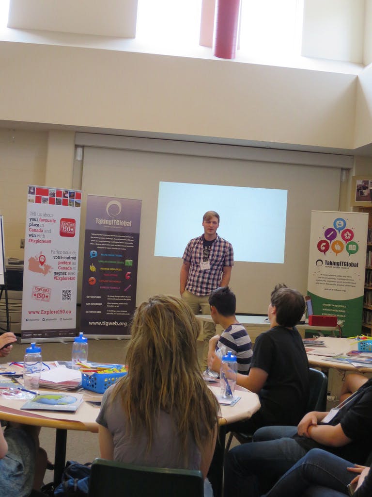 An educator stands at the front of the class, presenting to several students sitting around circular tables in the foreground. 