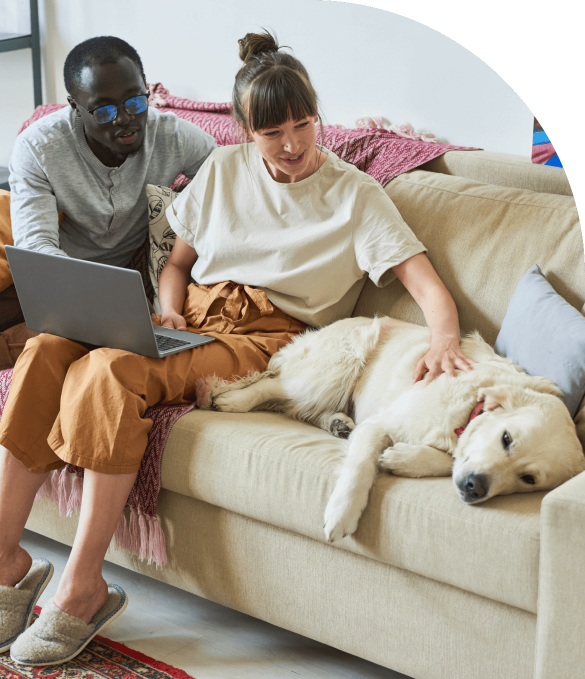 A photo of a couple sitting on their couch, sharing the screen of a laptop and petting their sleepy yellow lab