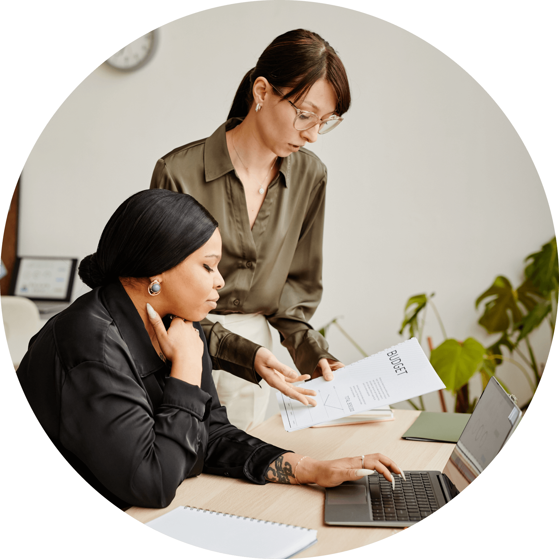 Circular photo of two women reviewing paperwork together in an office setting in front of a laptop