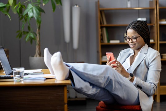 business woman reclines, with her socked feet up her desk, looking her cell phone and ignoring the work in front of her