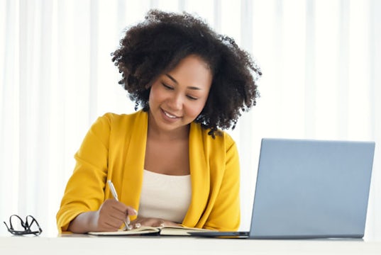 African-American entrepreneur sits at her laptop jotting down ideas in a notebook