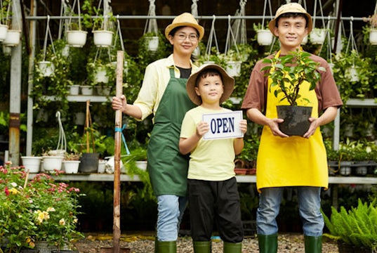 Family stands in their flower shop. The Dad holds a small plant and the son holds an open sign.
