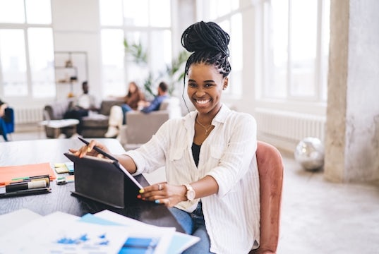 Young entrepreneur smiles and sits at her desk while working at a tablet