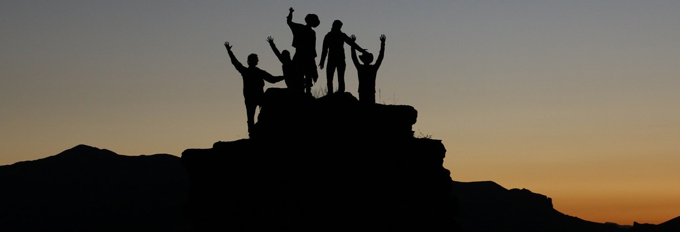A photo with a silhouette of a team of 5 people raising their hands standing on the top of a hill at dusk. 