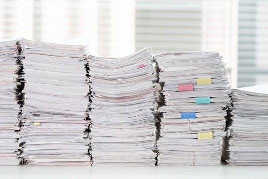 Stacks of paperwork are piled very high on a desk and blocking a window