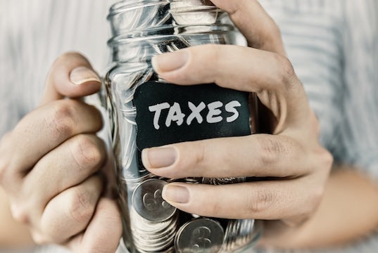 Hands tightly hold a jar labeled "taxes" that is filled with coins and dollars