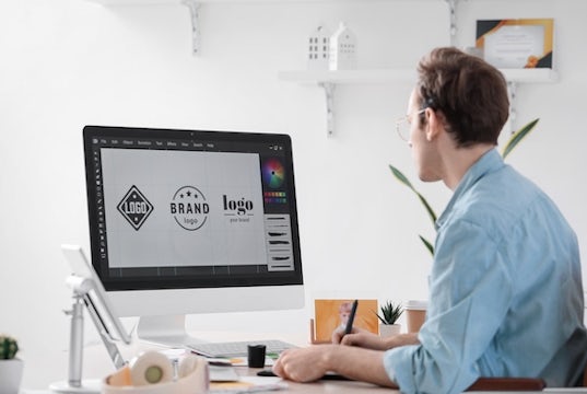 A man sits at his computer working on different logo design options on his screen 