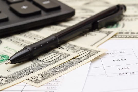 A pen sits next to a calculator with some dollar bills and a ledger