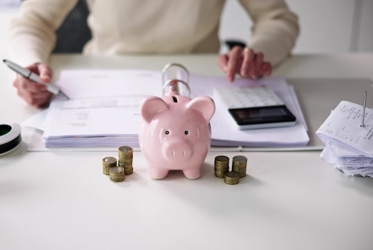 An aspiring business owner sits at a desk with a piggy bank, calculator, and ledger
