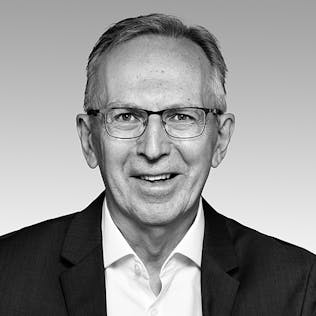 Ein lächelnder Mann mit Brille, weißem Hemd und schwarzem Sakko.