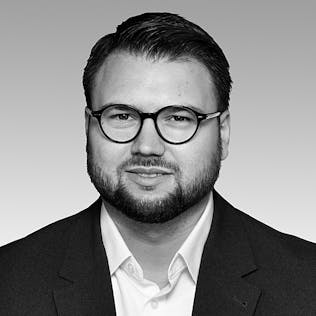 Ein lächelnder Mann mit Brille, weißem Hemd und schwarzem Sakko.