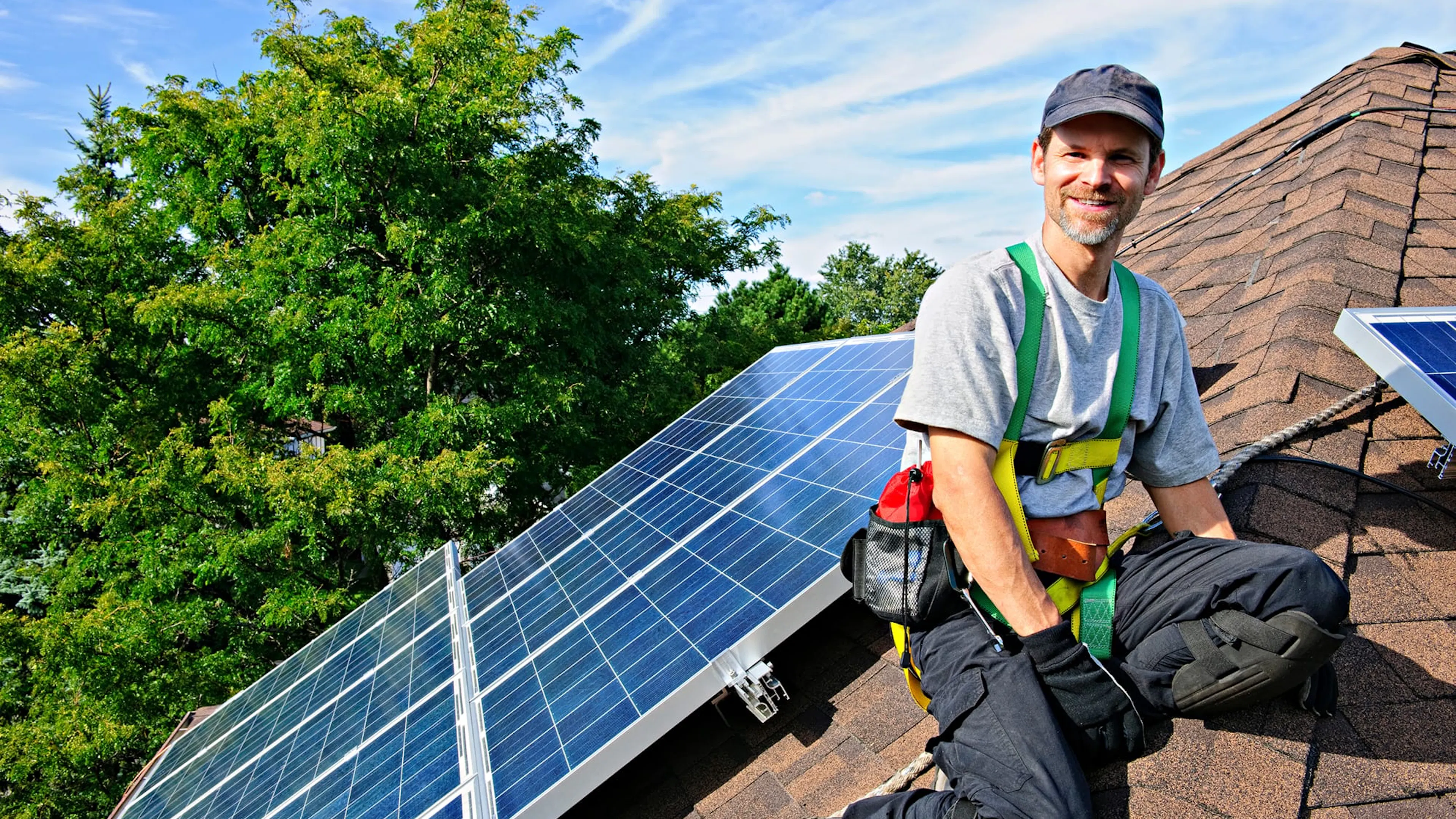 A solar installer sits on a home's roof, smiling, as he looks at the panels he just installed.