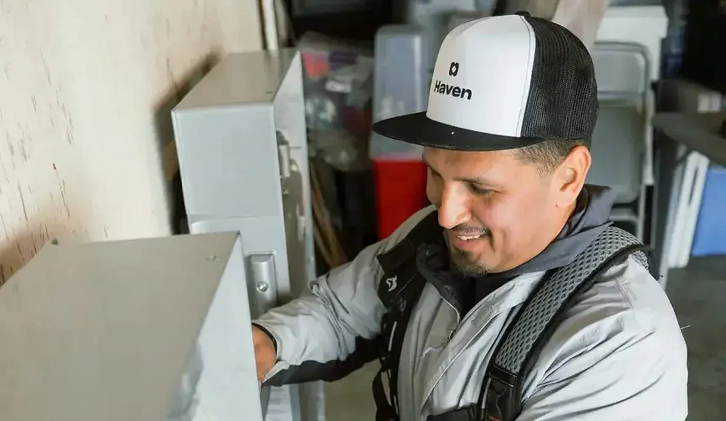 An electrician works on a home's meter socket, donning a hat branded with the company name, Haven.
