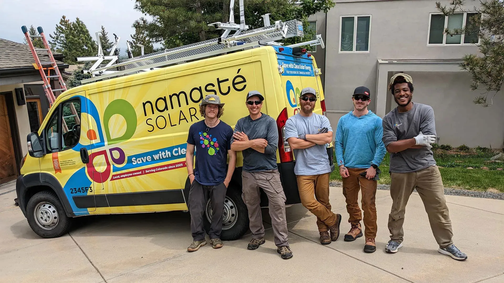 Crew members from Namaste Solar stand in a driveway next to their equipment truck, in front of a home with rooftop solar panels.