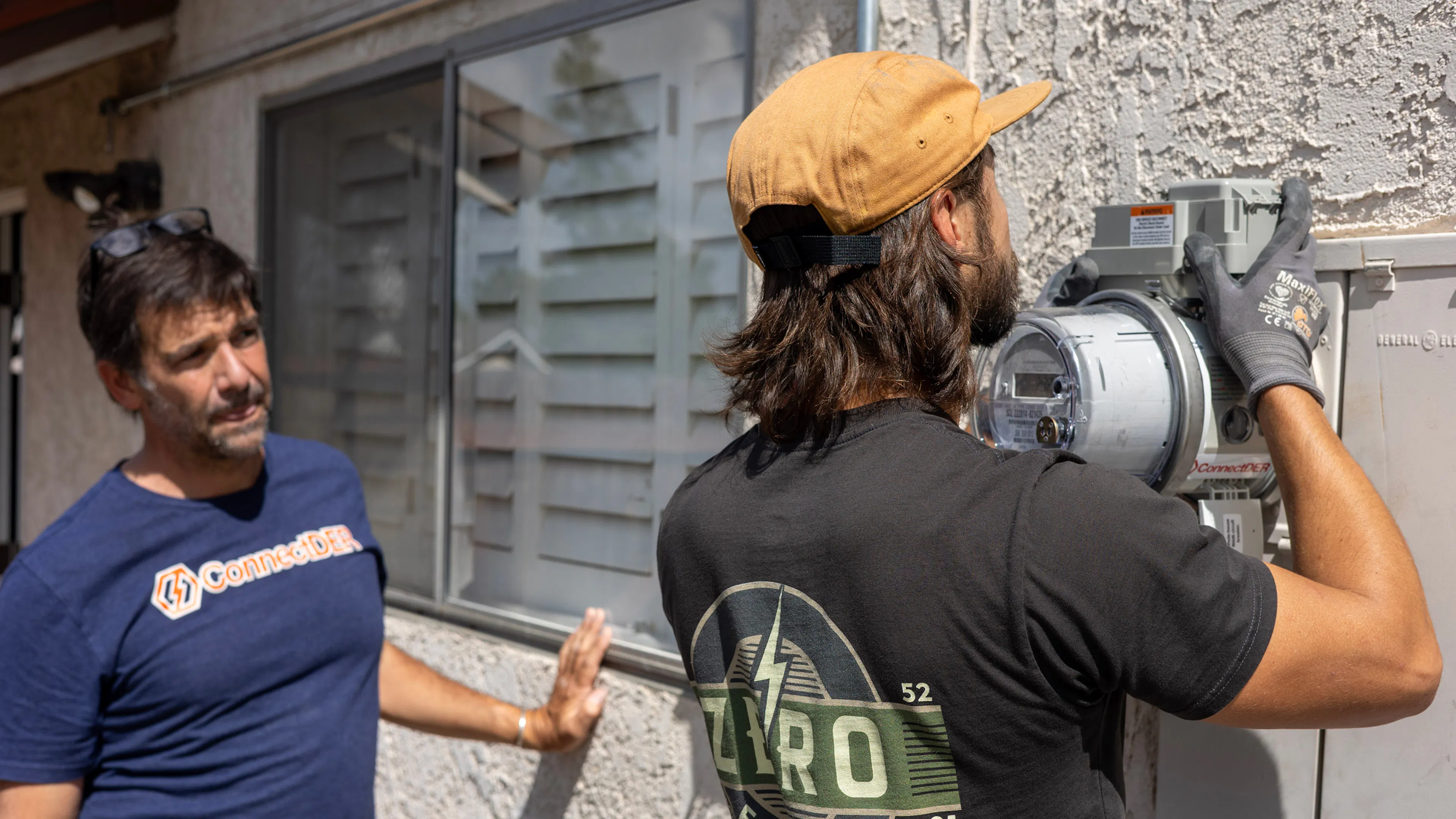 A crew member from Zero Electric installs the ConnectDER EV MSA into a meter socket on the side of a home, while a person in a ConnectDER tee shirt looks on.