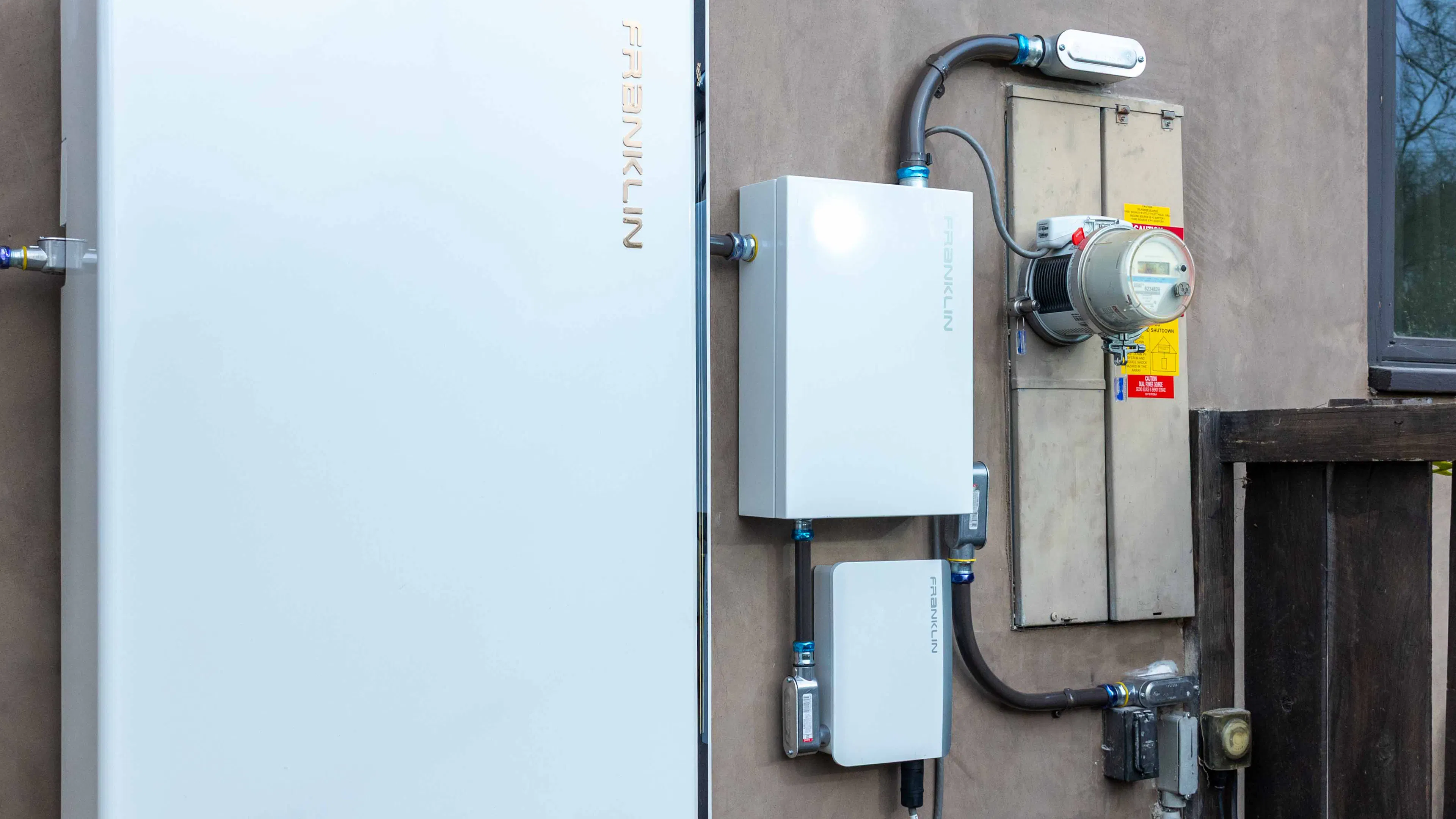 A battery storage setup on the side of a home showing the battery from FranklinWH, a meter adapter controller box, and the home's meter socket with a ConnectDER IslandDER meter collar plugged in.