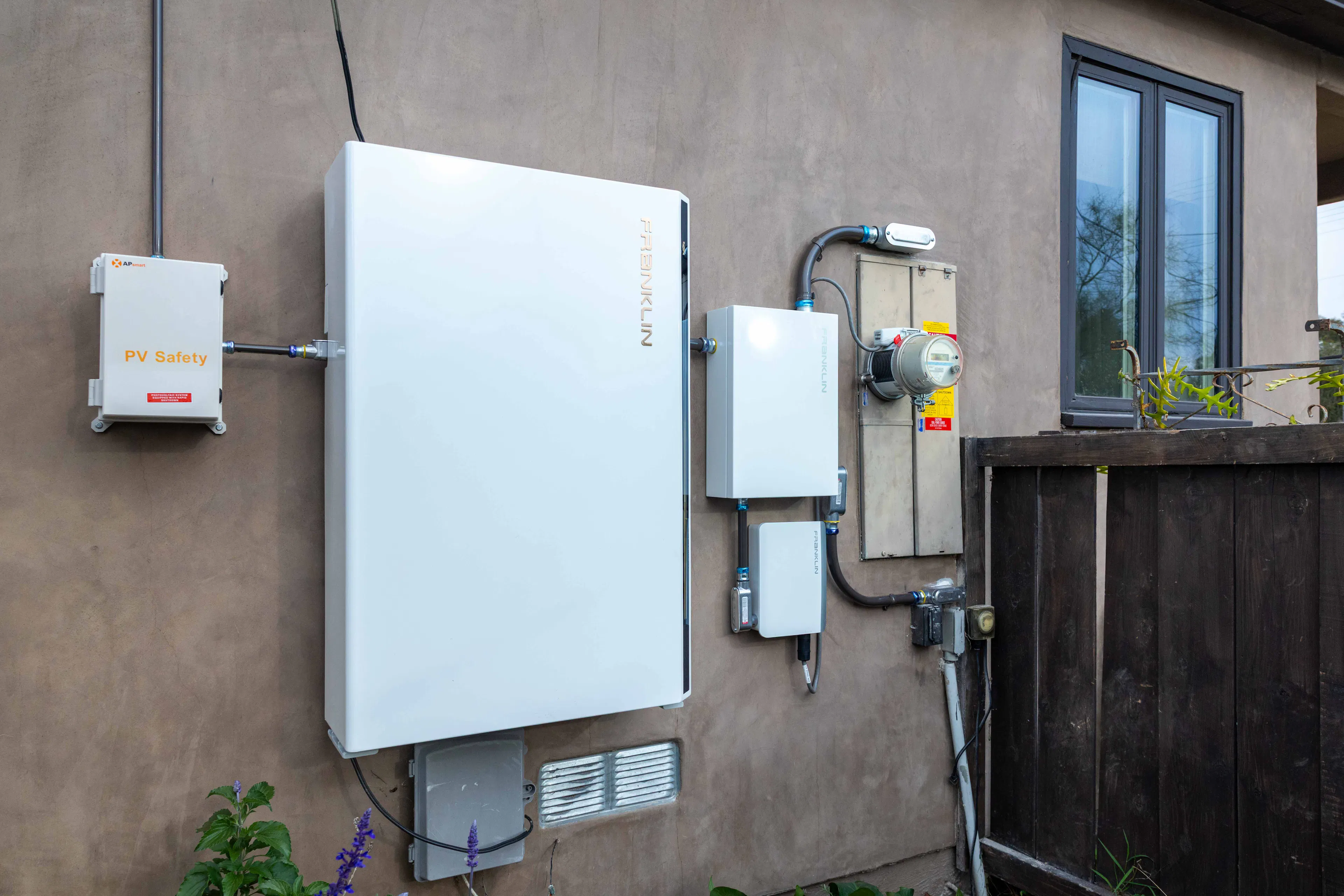 A battery storage setup on the side of a home showing the battery from FranklinWH, a meter adapter controller box, and the home's meter socket with a ConnectDER IslandDER meter collar plugged in.