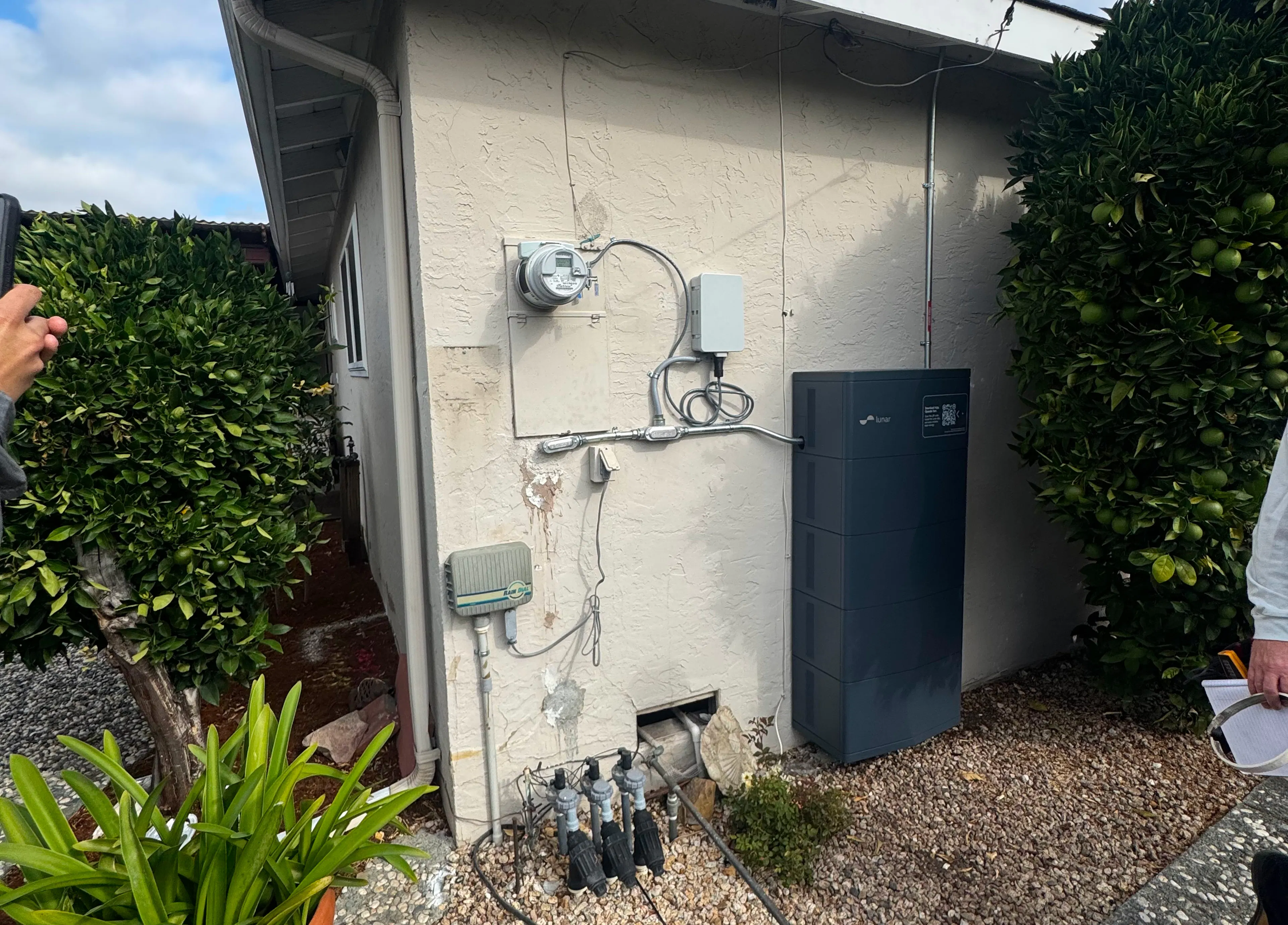 A battery storage setup on the side of a home showing the battery from Lunar Energy, a meter socket adapter controller box, and the home's meter socket with a ConnectDER IslandDER meter collar plugged in.