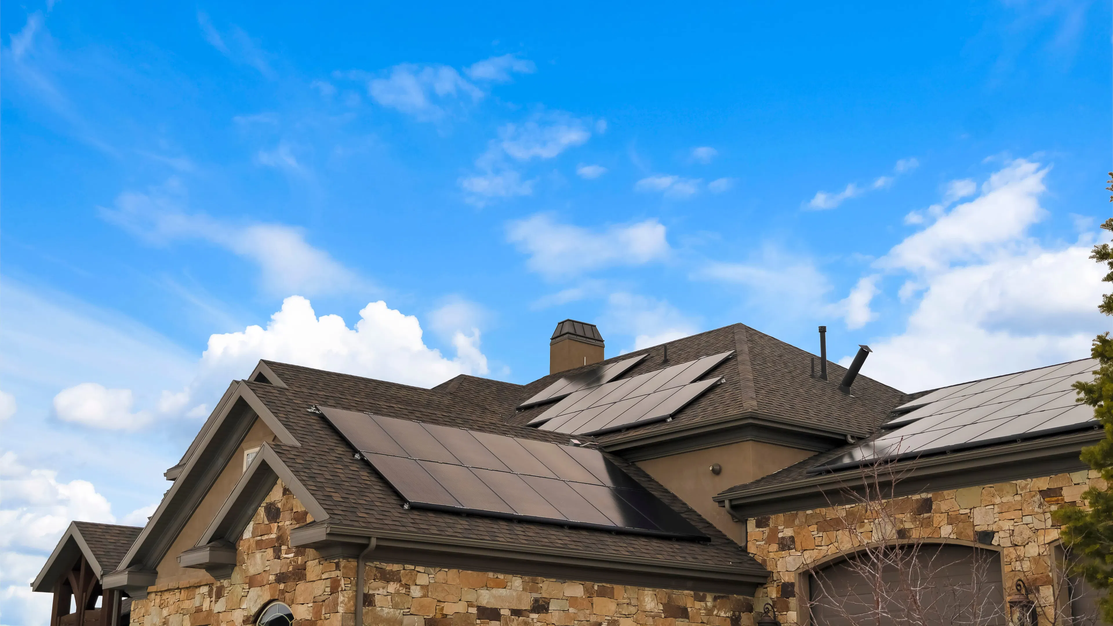 An upscale home with tan stone exterior siding and rooftop solar panels, underneath a bright blue sky.