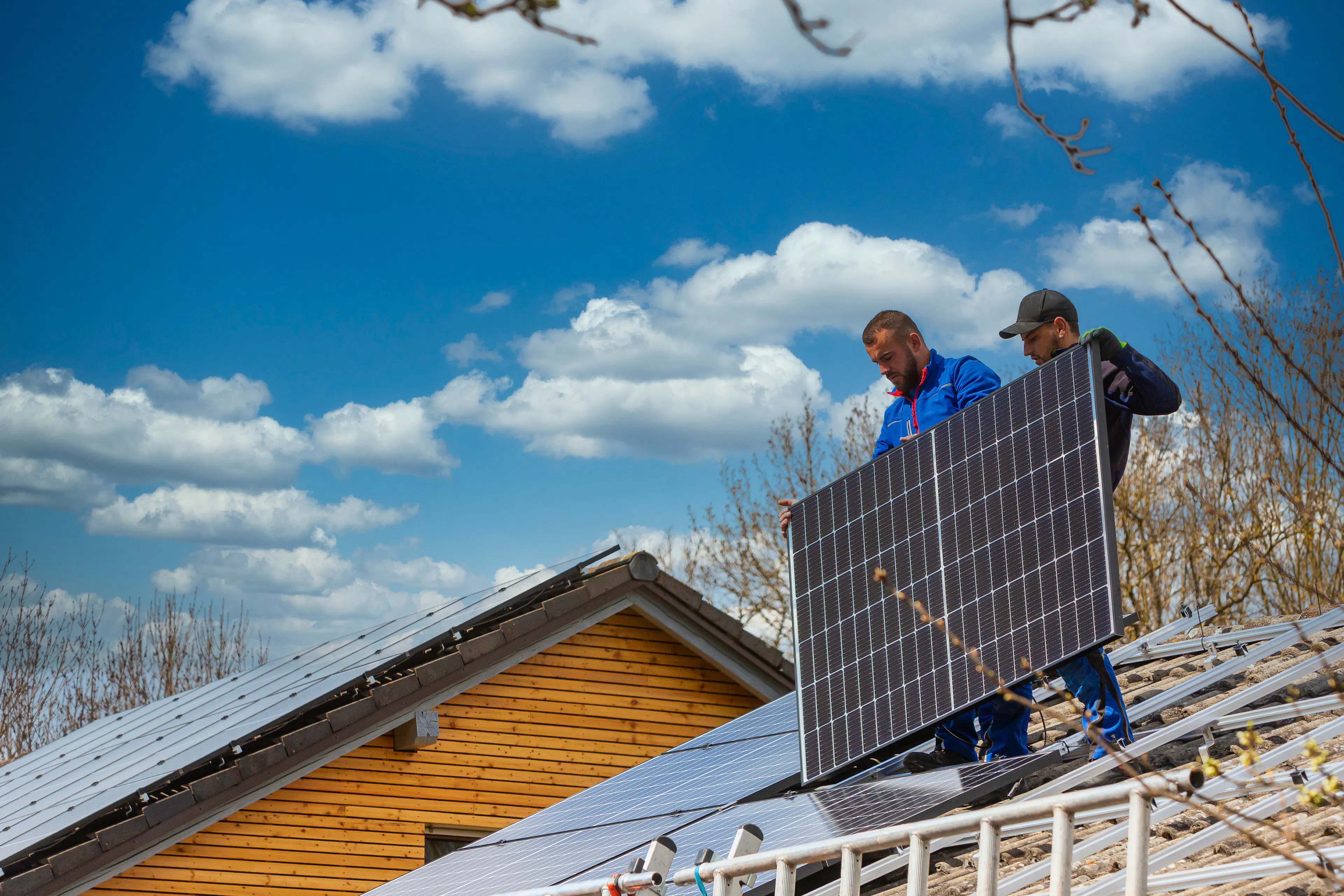 Solar installers on the roof of a home mounting a panel on a sunny day under blue skies.