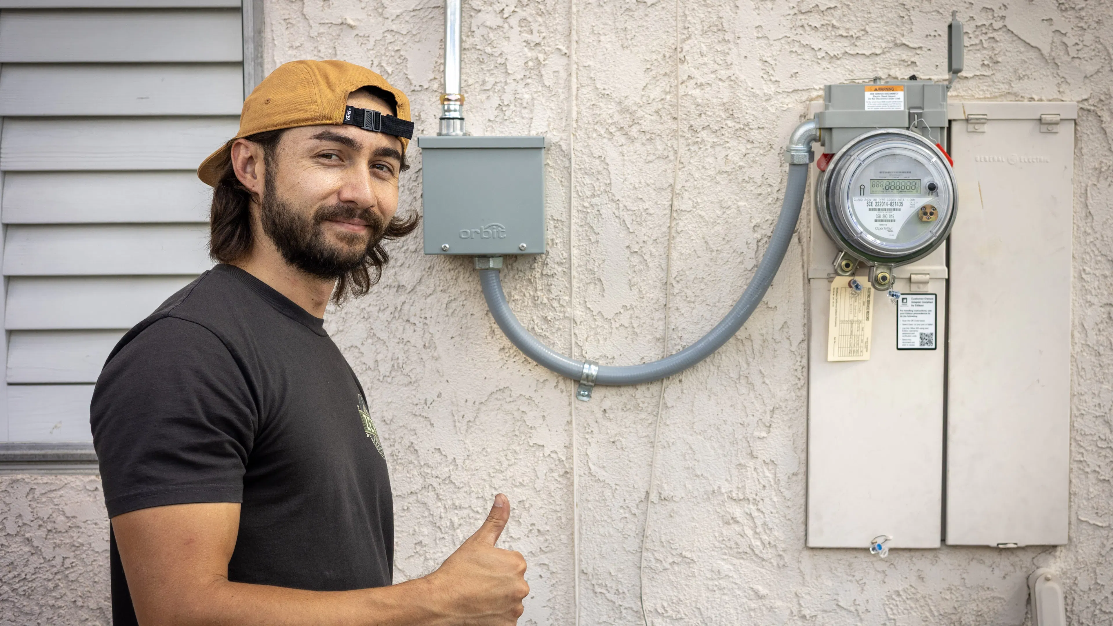An electrician standing outside a home at the meter socket with ConnectDER's EV Meter Socket Adapter installed, giving a thumbs up.