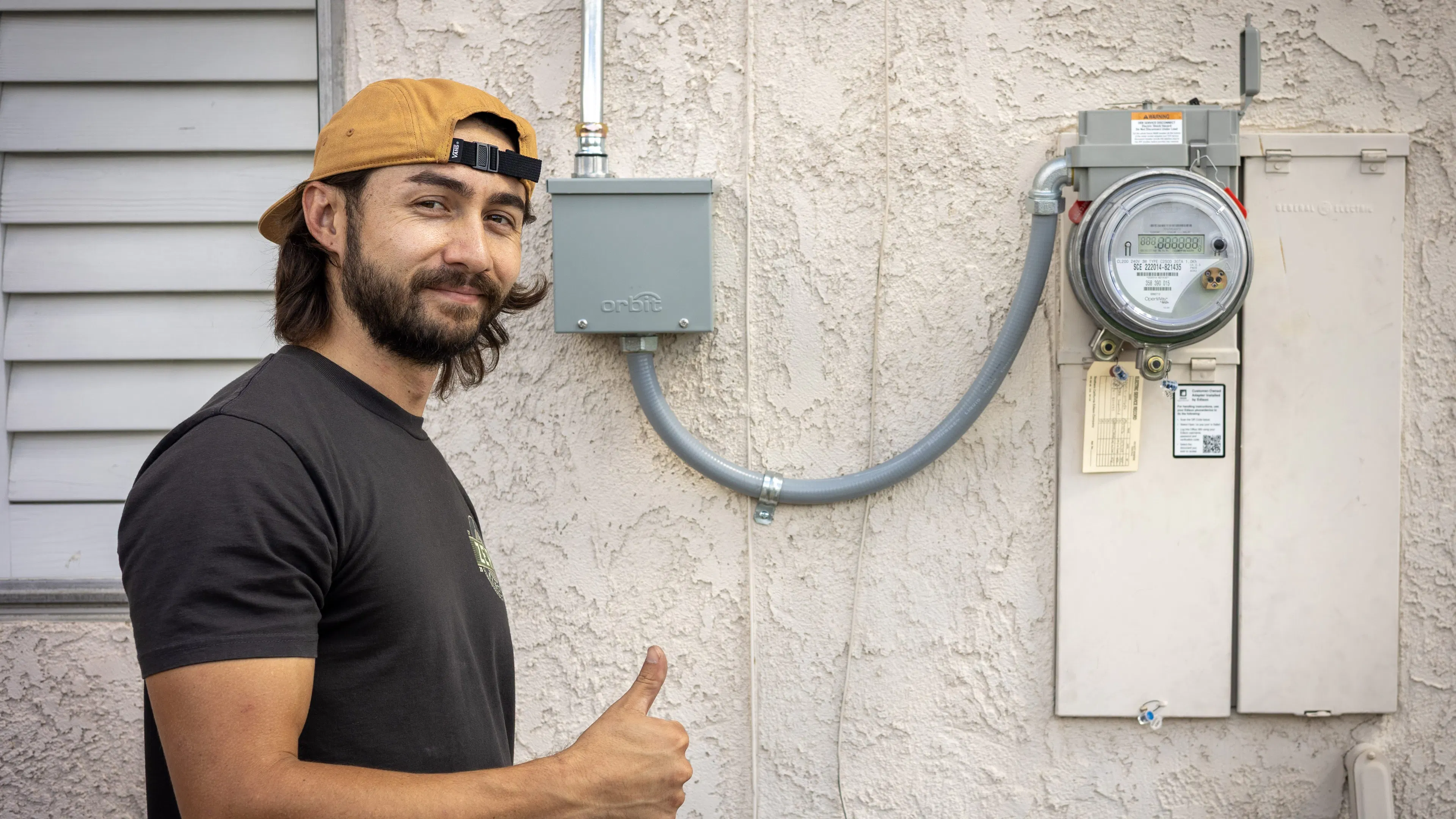 An electrician standing outside a home at the meter socket with ConnectDER's EV Meter Socket Adapter installed, giving a thumbs up.