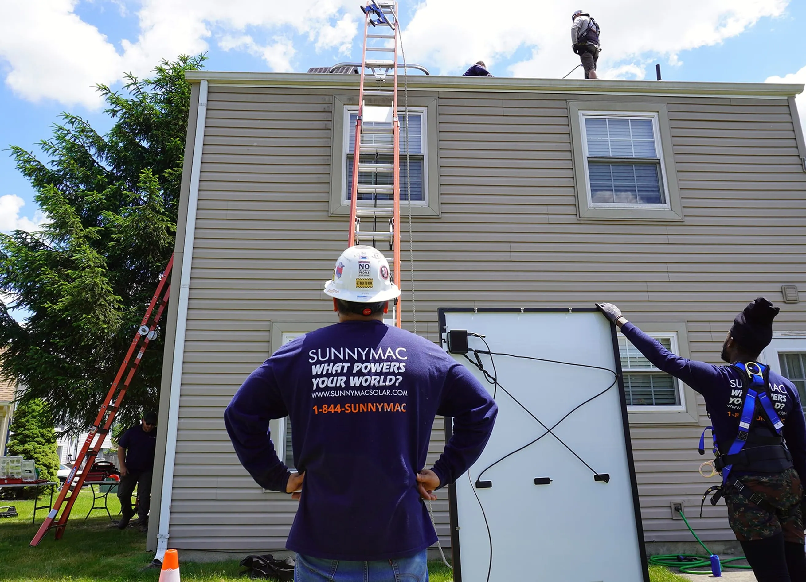 A crew member from Sunnymac stands in front of a home, watching his colleague bring a solar panel up to the roof of a house on a ladder.