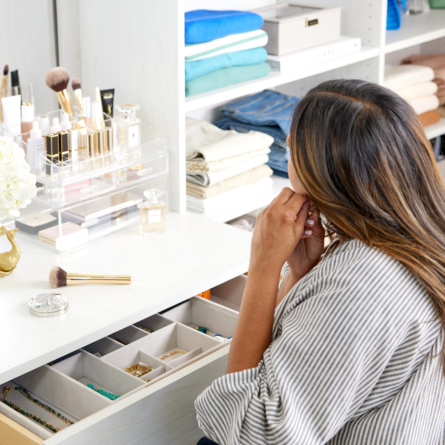 A woman putting on a pair of earrings, sitting at a vanity in her closet with a makeup organizer and drawer insert holding her jewelry.