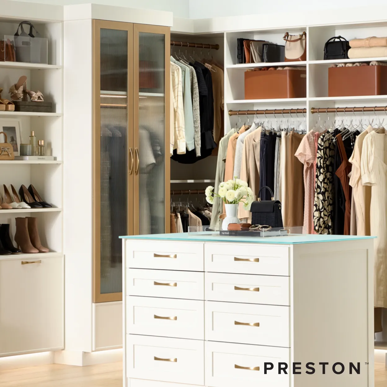 A white Preston custom closet with a center island of drawers, hanging clothes, and handbags on open shelves.