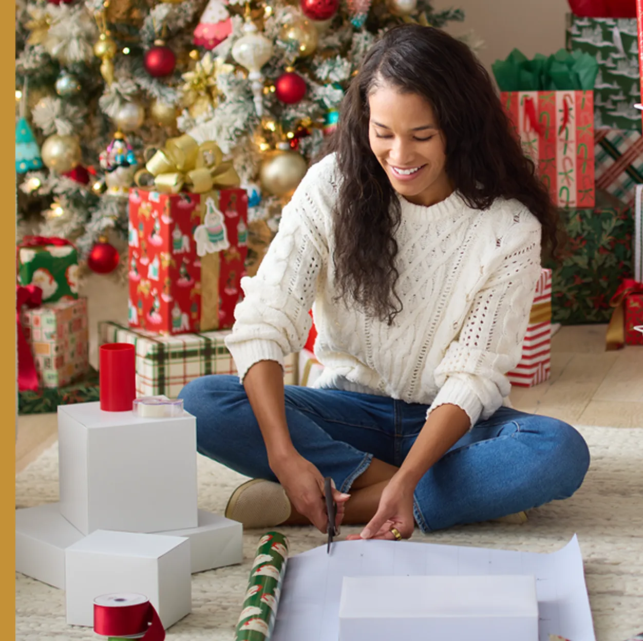 Woman sitting on the floor in front of a decorated Christmas tree, smiling as she cuts wrapping paper beside stacked white gift boxes, rolls of holiday wrap and red ribbon.