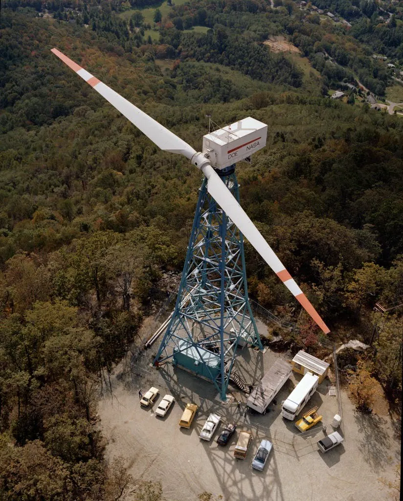 Aerial photograph of a large two-blade experimental turbine on a blue lattice tower in forest, vehicles parked at its base