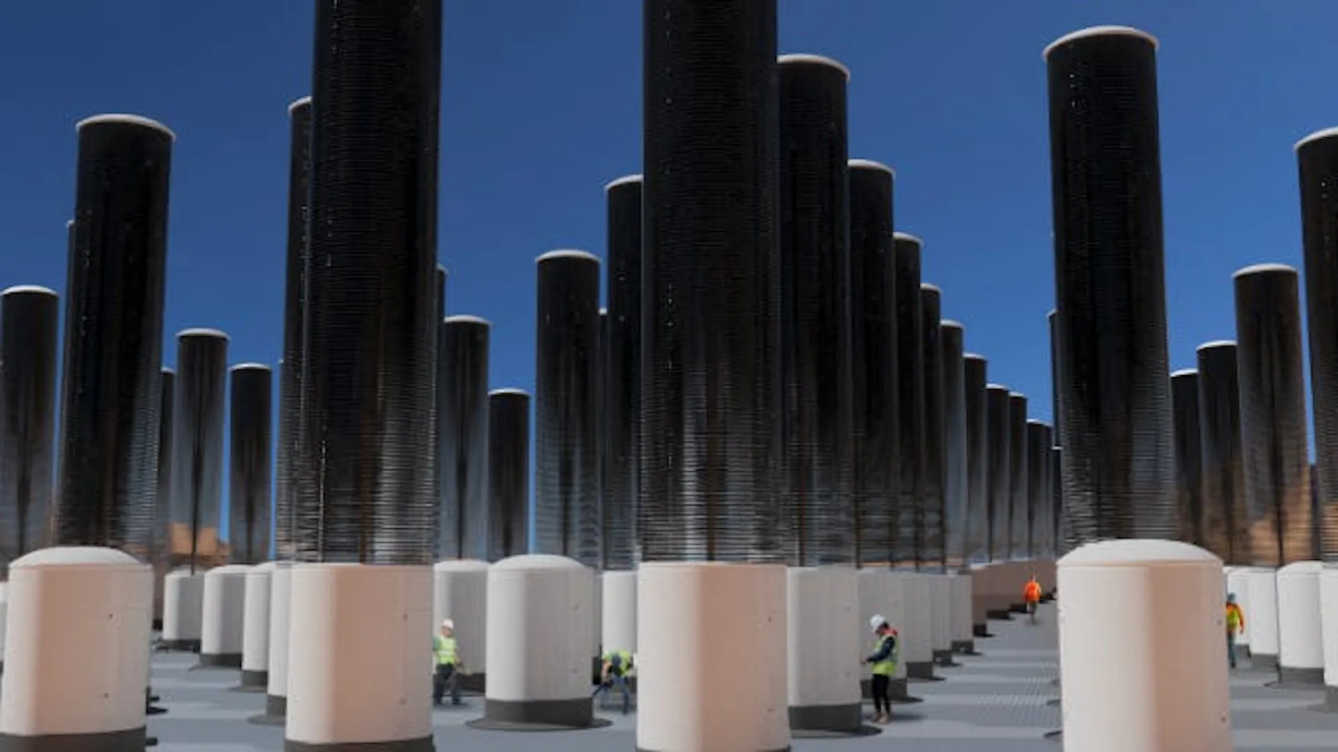 Photograph of tall cylindrical direct air capture towers in rows, workers walking between them