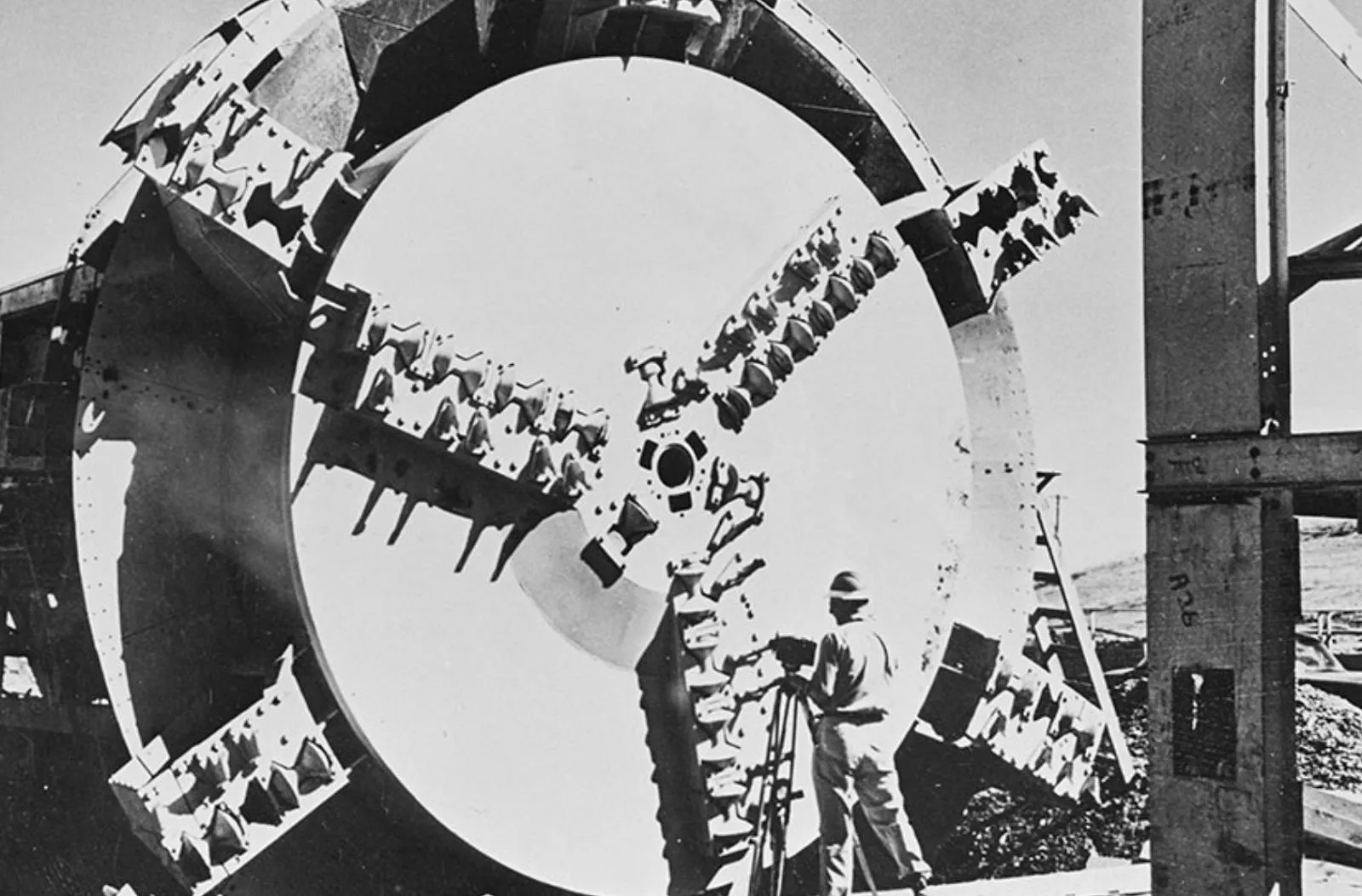 Black-and-white photograph of an early tunnel boring machine's cutting head, workers beside its ring of teeth