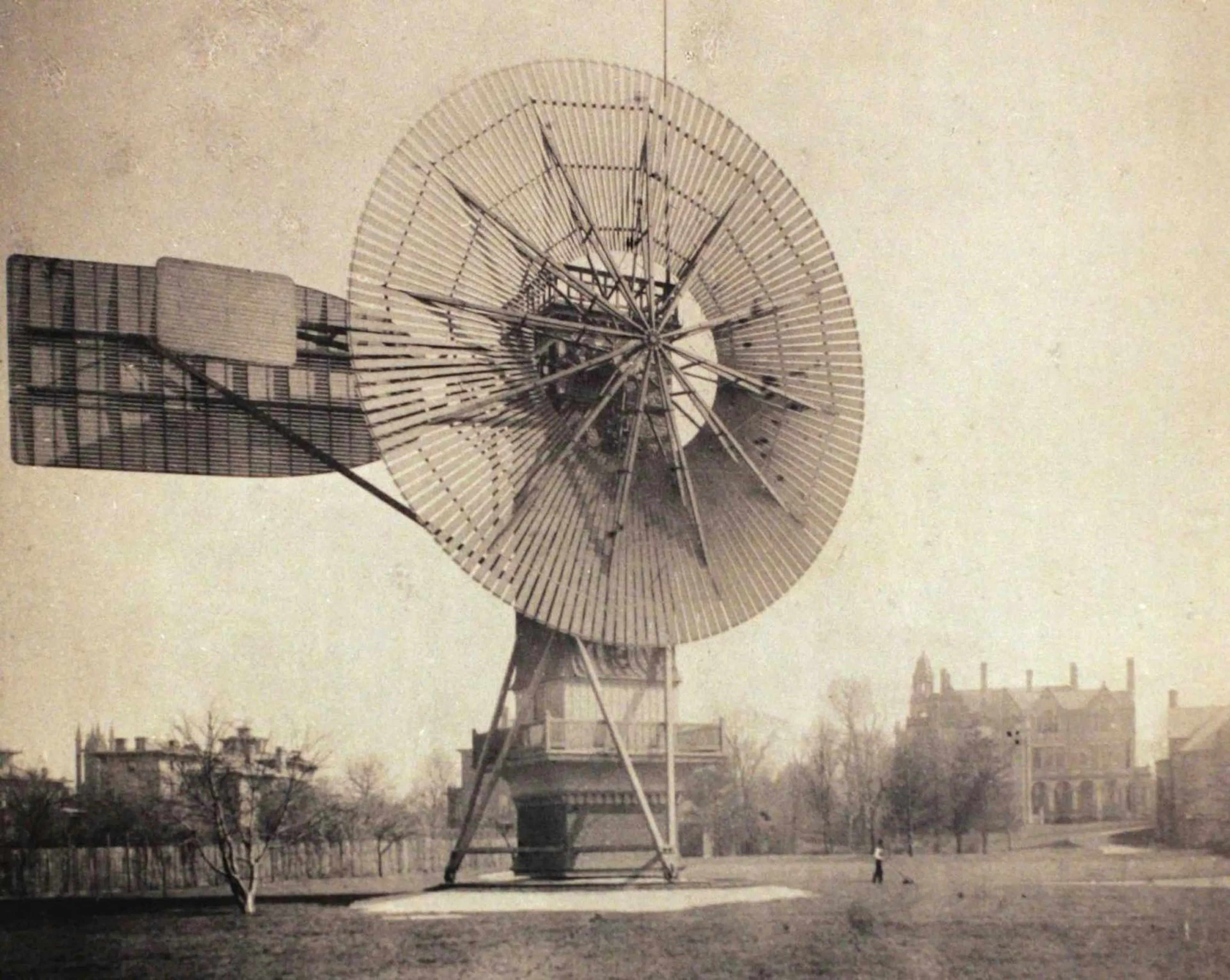 Sepia photograph of Charles Brush's multi-bladed wind dynamo, a large spoked rotor with tail vane on a timber frame, mansion grounds behind