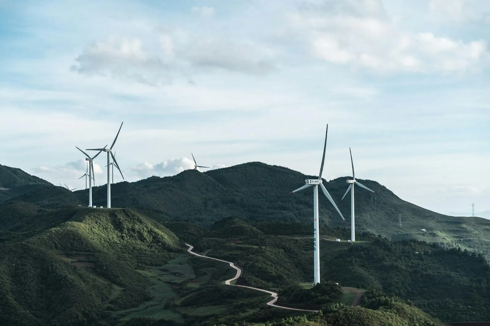 Modern white wind turbines spaced along green forested ridges above a winding road