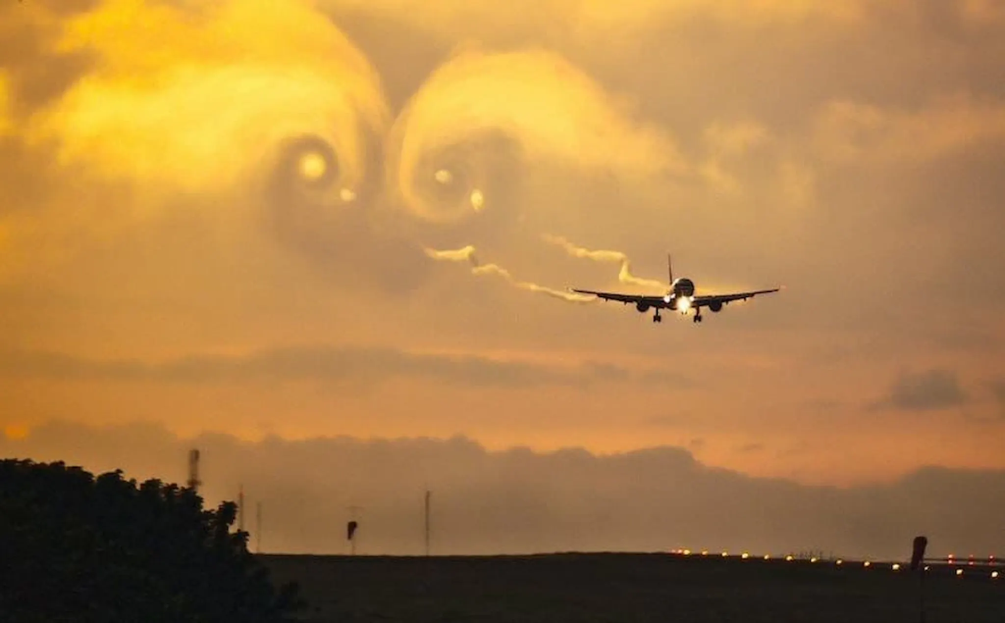 Photograph of an airliner on approach at sunset, its wingtip vortices carved visibly into golden cloud