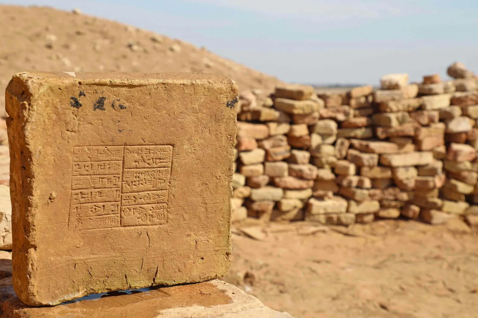 Photograph of a mud brick stamped with cuneiform, stacked bricks and desert behind