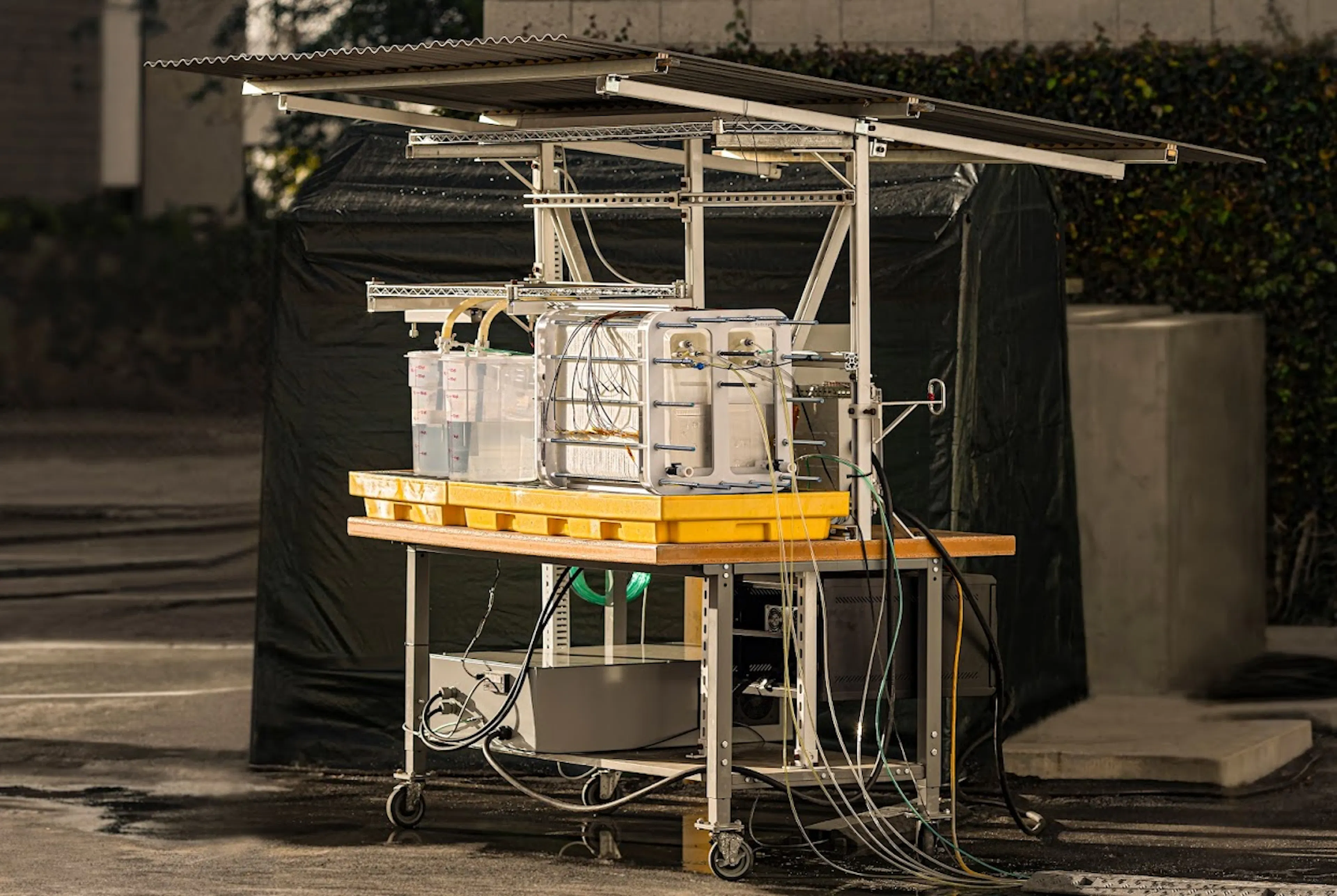 A prototype Terraform electrolyzer test rig on a wheeled cart under a canopy of solar panels, with electrolyzer cells, tanks, and cabling